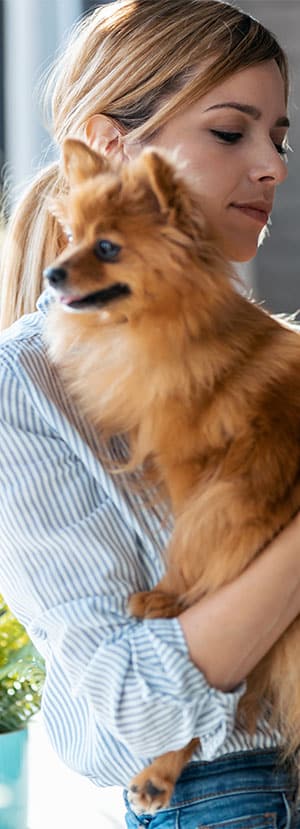 A woman with blonde hair in a ponytail, wearing a striped shirt, holds a small brown fluffy dog. The woman's eyes are closed, and she appears to be standing in a softly lit indoor setting, reminiscent of a cozy veterinarian’s office.